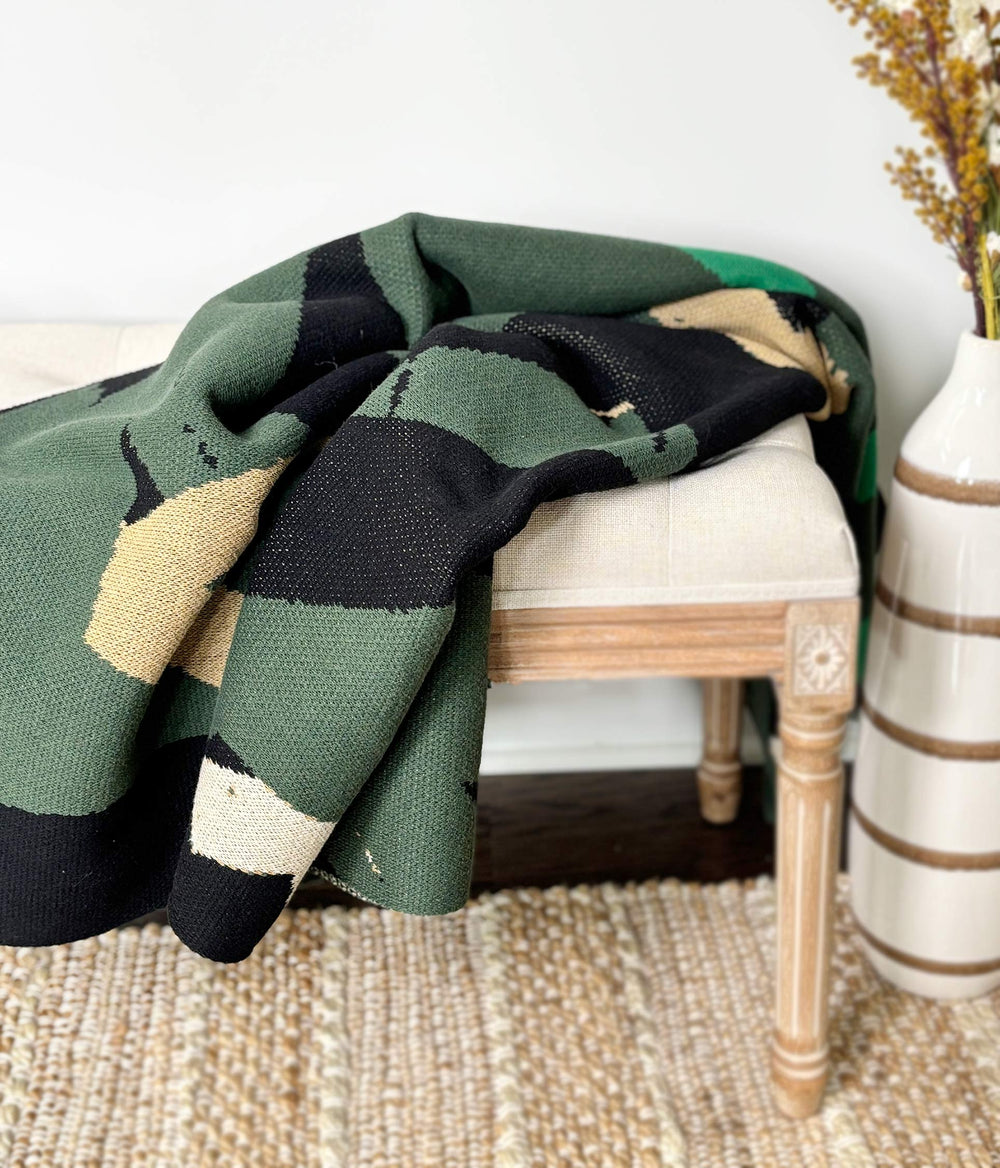 Green and black patterned blanket draped over a wooden chair with a vase in the background.