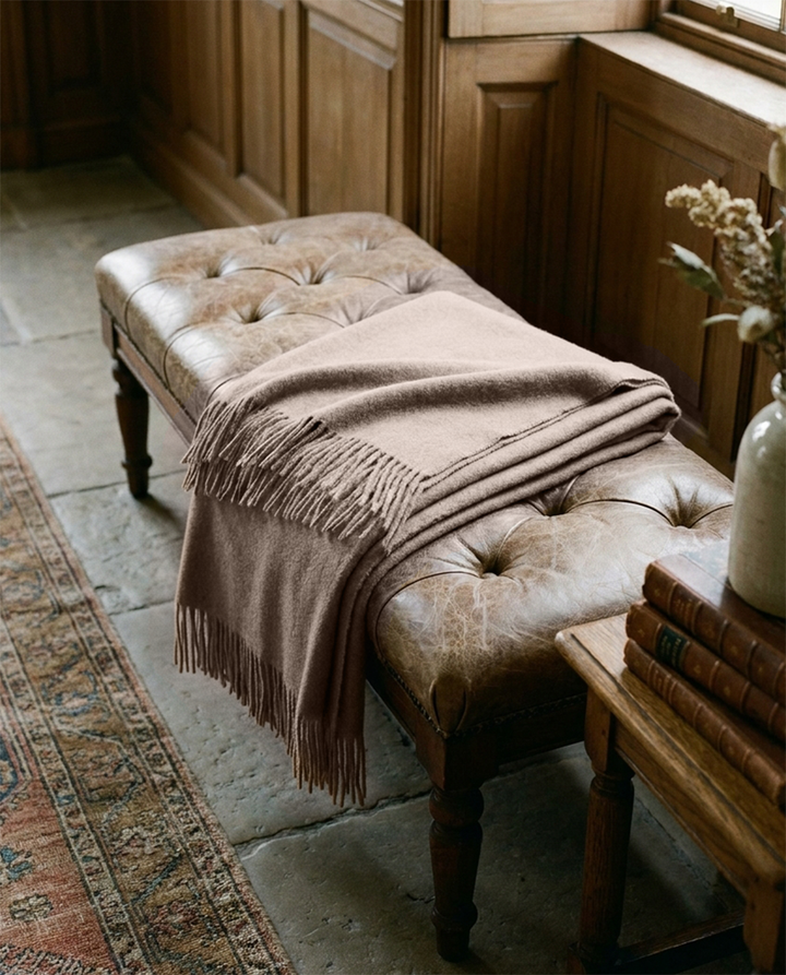 A taupe-brown knitted throw blanket with fringe is draped over a tufted leather bench in a rustic room with wood paneling and stone flooring.