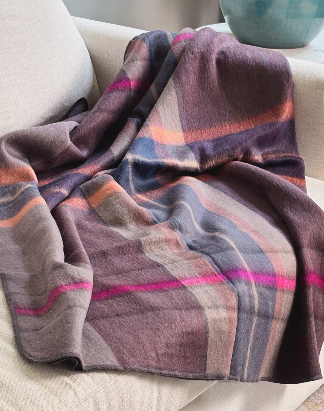 A soft plaid throw blanket draped over a light beige upholstered armchair, featuring muted purple, dusty rose, orange, and gray stripes with bright pink accents.