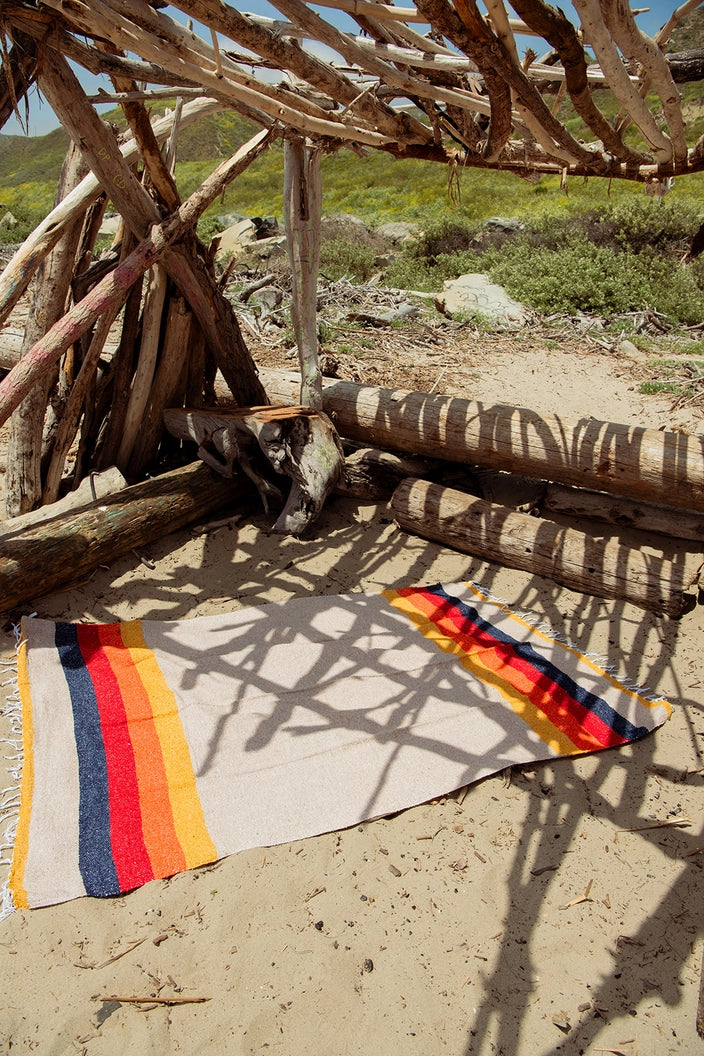 Blanket with colorful stripe patterns on a sandy ground under a wooden shelter.