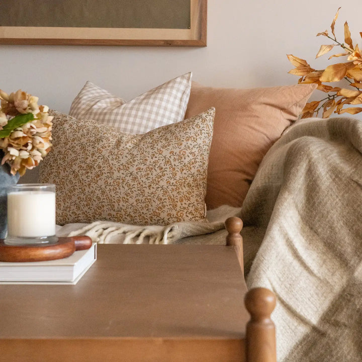 A cozy bedroom or living space scene with a wooden side table in the foreground, a beige throw blanket draped over a sofa, patterned and solid accent pillows, a candle, flowers, and a framed artwork on a light wall.