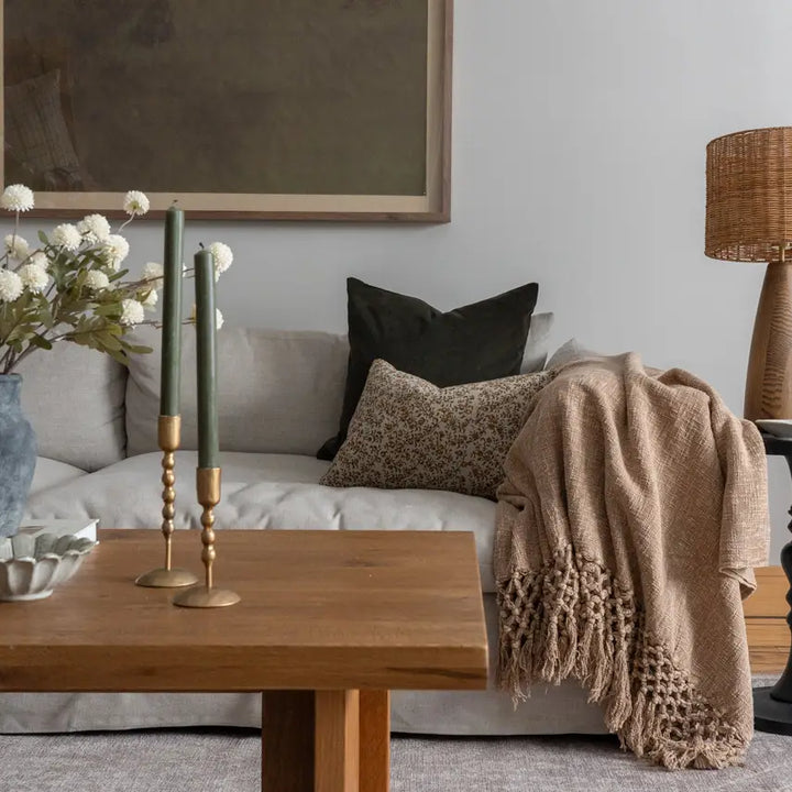 A cozy living room scene with a light beige sofa, a chunky knit tan throw blanket draped over the arm, dark green and leopard-print pillows, a wooden coffee table, and a vase of white flowers.